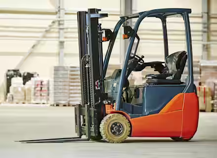A red and black forklift is parked indoors on a concrete floor. In the background, there are stacks of boxes and pallets against a large industrial wall. The warehouse setting is well-lit.