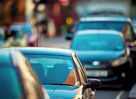 A row of parked cars lines a city street, with the foreground car in sharp focus and the background cars and buildings blurred.