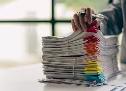A hand holding a pen is seen organizing a tall stack of paper files, which are secured with rubber bands and sorted using multicolored tabs. The scene is set on a white desk with a blurred window in the background.