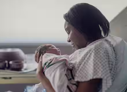A mother in a hospital gown gently cradles her newborn baby in a hospital bed. Both mother and baby are looking at each other tenderly. The baby is wrapped in a blanket. There are hospital items in the background on a tray.