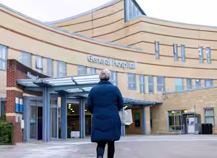 A person in a blue coat and holding a white bag walks toward the entrance of a modern building labeled "General Hospital" on a cloudy day.