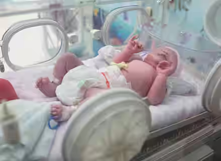 A newborn baby lies in an incubator, wearing a diaper and a cap. The baby is covered with a blanket, and medical equipment surrounds the incubator, indicating a hospital setting.