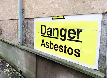 A yellow and white sign on a boarded-up building wall reads "Danger Asbestos," warning of hazardous materials inside.