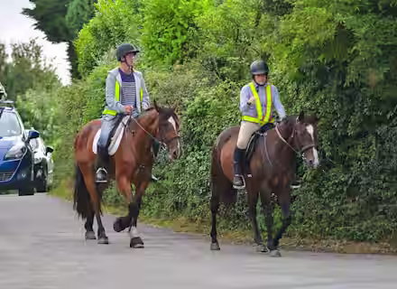 Two people, wearing helmets and reflective vests, are riding brown horses along a quiet, paved road bordered by lush green hedges. In the background, a parked blue car is partially visible. The riders appear to be relaxed and enjoying their leisurely ride.
