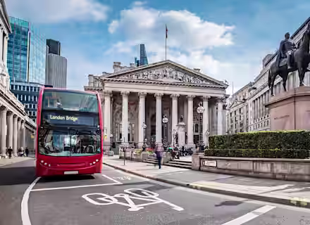 A red double-decker bus with the destination "London Bridge" displayed on its sign passes in front of the Royal Exchange in London. Modern office buildings and a statue of a man on horseback are visible in the background under a partly cloudy sky.