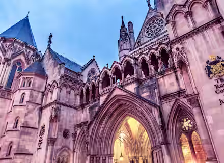 Image of the exterior of The Royal Courts of Justice in London, featuring Gothic architecture with intricate arches, pointed spires, and detailed carvings. The building's name is visibly inscribed on the wall along with a crest, illuminated by warm lighting from within.