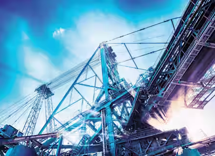 A towering industrial structure with cranes and metal beams is shown against a bright blue sky. The scene depicts a construction or manufacturing facility, with beams of light creating an energized atmosphere. Cloudy elements and sun rays filter through the industrial setting.