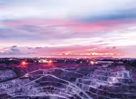 A large open-pit mine with tiered levels, illuminated by several bright lights, stretches across the foreground under a colorful sky at sunset. In the distance, city lights twinkle, blending with the vibrant hues of the sky.