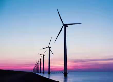 A row of wind turbines stands tall against a vibrant sunset backdrop over a body of water. The sky is painted with hues of pink, purple, and blue as the day transitions into evening. The turbines are aligned in perspective, stretching into the distance.