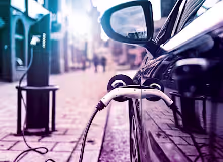 A close-up shot of an electric vehicle being charged at a charging station on a city street. The car's side mirror and charging port are prominently in view, with the plug connected. The background is slightly blurred, showing buildings and streetlights.
