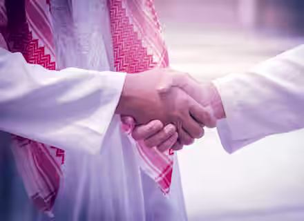 Close-up of two people wearing traditional Middle Eastern clothing shaking hands. Both individuals are dressed in white garments with one person wearing a red and white checkered keffiyeh. The background is blurred, emphasizing the handshake.