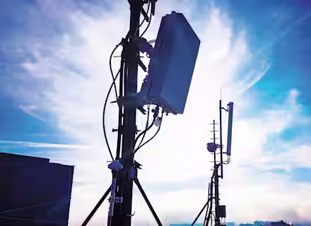 Silhouettes of telecommunications towers with various antennas and equipment are seen against a bright, cloudy sky at sunset or sunrise. The scene is set atop a building in an urban area.