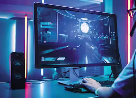 A person wearing glasses and headphones is sitting at a desk, playing a futuristic video game on a large monitor. The room is illuminated with blue and pink neon lights, and speakers are placed on both sides of the monitor.