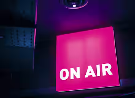 A brightly illuminated pink "ON AIR" sign is mounted on a dark wall, signaling that a live broadcast is in progress. The surrounding area is dimly lit, highlighting the glow from the sign.