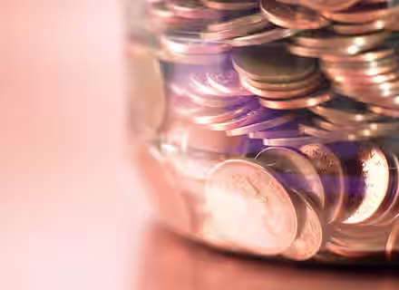 A glass jar filled with copper coins, mostly pennies, displayed against a softly blurred background. The coins reflect light, showing their shiny surfaces and engraved details. The jar is nearly full, with coins tightly packed inside.