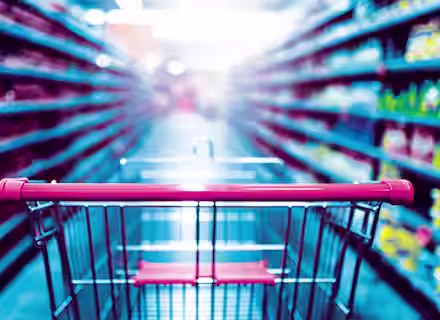 A shopping cart with a red handle is in focus in the foreground of a brightly lit supermarket aisle, surrounded by shelves stocked with a variety of products. The background is blurred, emphasizing the perspective from the shopper's point of view.