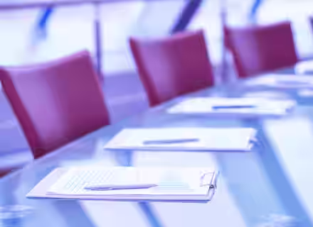 A glass conference table is set with clipboards, pens, and papers. Red chairs are arranged around the table in the background, with a soft focus on the modern, light-filled space. The overall tone of the image is cool, with blue and purple hues.
