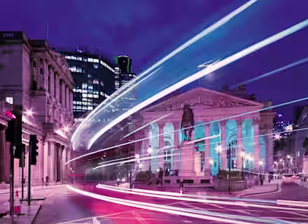 A nighttime cityscape featuring a neoclassical building with columns, illuminated by colorful lights. Light trails from passing vehicles create dynamic streaks, and modern skyscrapers are visible in the background, blending historic and contemporary architecture.