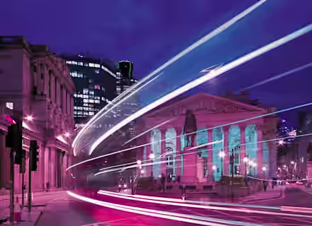 A nighttime cityscape featuring the Royal Exchange in London, illuminated with pink and blue lights. Light trails from passing vehicles create dynamic streaks in the foreground. Surrounding buildings are also visible under a deep blue and purple sky.