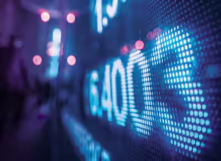 A close-up view of a digital screen displaying financial data, including numbers like 1.389 and 4003, with a purple and blue hue. The background is blurred, showing a soft-focus interior setting and glowing orange lights.