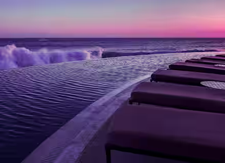 Infinity pool overlooking the ocean at sunset. Waves crash gently on the edge, and the sky is painted in hues of pink and purple. Several poolside lounge chairs are lined up on the right, suggesting a serene and luxurious setting.