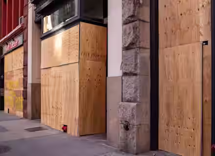 A city street view showing multiple storefronts with windows and doors covered in wooden boards. The building on the left has red paint, while the others have a combination of stone and brick facades. The area appears calm with no people visible.