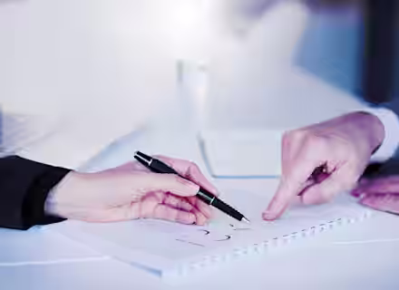 Two people, one in a dark suit and the other in a light suit, are seated at a table. One person is holding a pen and pointing to a document on the table, while the other person is pointing at the same document. The scene suggests a discussion or review of the document.