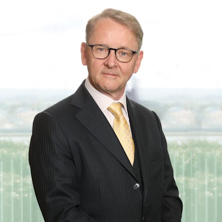 A man with short, light hair and glasses stands against a light-colored background. He is wearing a striped, dark suit with a light yellow tie and a light-colored shirt, looking directly at the camera with a neutral expression.