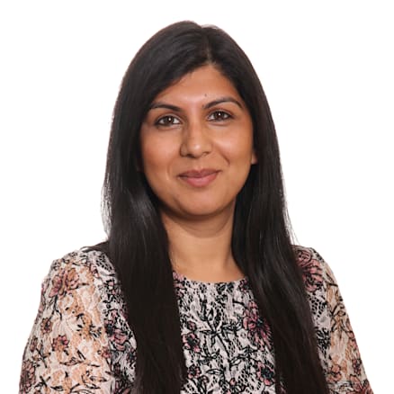 A woman with long, straight black hair is smiling. She is wearing a short-sleeve blouse with a floral pattern of black, pink, and beige shades. The background is plain white.