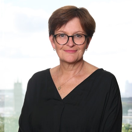 A woman with short brown hair and glasses stands in front of a large window with a cityscape view. She wears a black, long-sleeved top and a gold-colored necklace. She has a neutral expression and her hands are clasped in front of her.