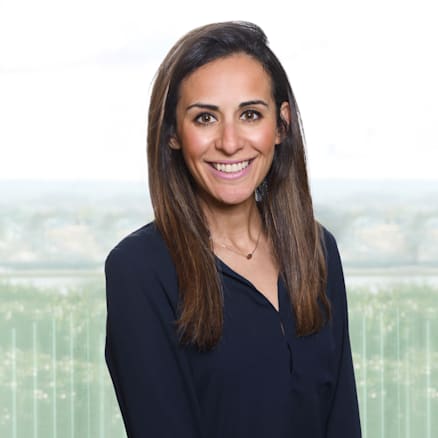 A woman with long, straight brown hair is smiling. She is wearing a dark blue blouse and stands with her hands in front of her. The background is a blurred outdoor scene with natural light filtering through.