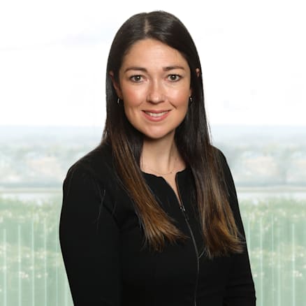 A woman with long, straight brown hair and a light complexion is smiling at the camera. She is wearing a black long-sleeve top and standing in front of a blurred background featuring a window view.