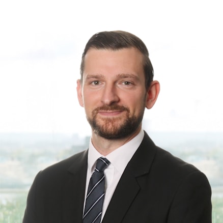 A man with short dark hair and a trimmed beard is wearing a black suit, white dress shirt, and a striped tie. He is posing for a professional portrait in front of a blurred outdoor background.