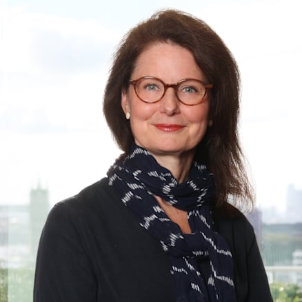 A woman with shoulder-length dark hair and glasses is smiling slightly. She is wearing a dark long-sleeve top and a patterned scarf. The background shows a cityscape with tall buildings under a cloudy sky.