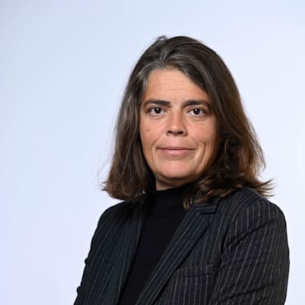 A person with shoulder-length dark hair stands against a plain white background, arms crossed, wearing a dark pinstripe blazer and black top, looking at the camera with a neutral expression.