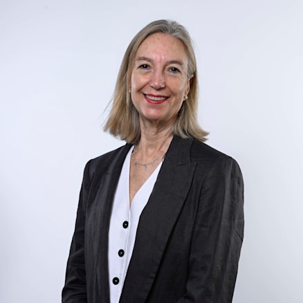 A woman with shoulder-length blonde hair smiles at the camera. She is wearing a dark blazer over a white blouse with a V-neck and buttons, standing in front of a plain white background. Her hands are clasped in front of her.