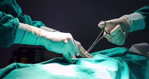 Two surgeons in green surgical gowns and gloves perform a procedure on a patient, using surgical instruments under focused lighting.