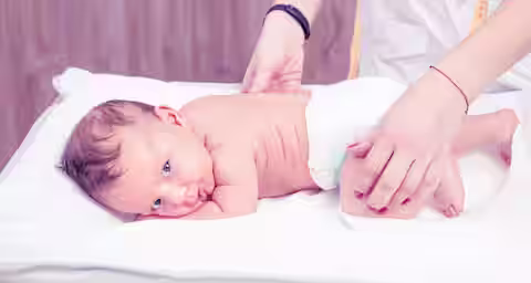 A baby lies on their stomach on a soft white surface, wearing a diaper. An adult's hands, one with a tape measure, gently touch the baby's back and leg. The background is softly blurred, giving a warm and calm atmosphere.