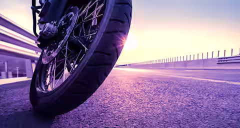 Close-up of a motorcycle wheel on an empty road at sunset. The sun is low on the horizon, casting a warm glow on the road and the motorcycle tire. The scene conveys a sense of freedom and adventure.