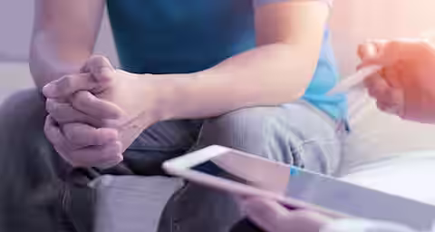 A person in a blue shirt and jeans sits with their hands clasped together on their lap, facing another person holding a digital tablet and a stylus. They appear to be engaged in a conversation or counseling session in a softly lit environment.