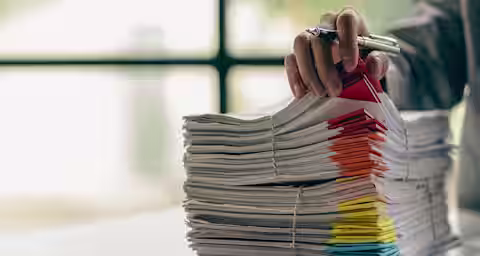 A hand holding a pen is seen organizing a tall stack of paper files, which are secured with rubber bands and sorted using multicolored tabs. The scene is set on a white desk with a blurred window in the background.