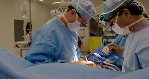 Two surgeons in blue surgical gowns and masks perform a procedure in a sterile operating room. One uses a head-mounted light for better visibility. Medical equipment surrounds them, and an overhead surgical light is illuminated, providing ample lighting.