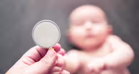A close-up of a hand holding a stethoscope near a baby lying on a dark surface. The baby is out of focus and appears to be reaching toward the stethoscope. The stethoscope has a yellow tube and a circular diaphragm.