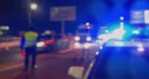 Blurred image of a nighttime scene with several cars on the road, flashing blue and red lights from police vehicles, and a person in a reflective vest standing by the roadside. The atmosphere suggests an ongoing emergency or traffic stop.