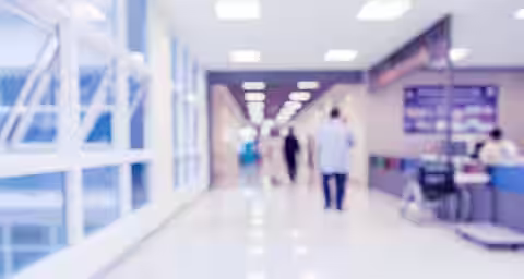 A blurred image of a hospital corridor with bright lighting. People are walking in the distance, and a wheelchair is positioned near a counter on the right side. Large windows on the left let in natural light. Tables with information are on the right side of the hallway.