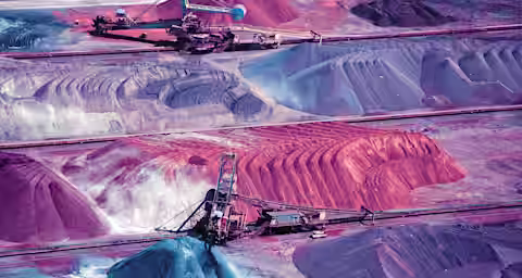 An aerial view of a large mining site with various piles of extracted materials. Piles range in color from grey and blue to red and orange. Heavy machinery is visible extracting and moving materials across the site. The terrain appears segmented with tracks.