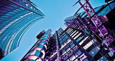 A low-angle view of modern high-rise buildings against a clear blue sky. The structures feature a mix of glass, steel, and vibrant purple lighting, showcasing architectural details with a futuristic appearance.