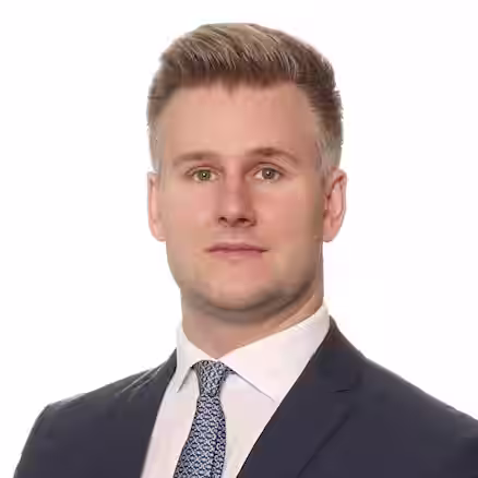 A professionally dressed man with short, neatly styled hair is seen from the shoulders up. He is wearing a navy blue suit, a white dress shirt, and a patterned tie. He has a neutral expression and is posed against a plain white background.