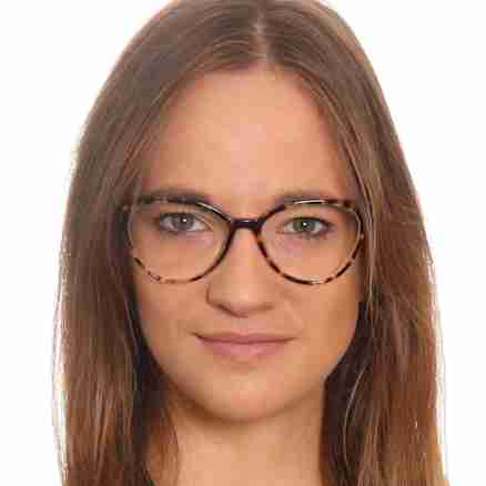 A woman with long light brown hair and glasses is posing in front of a white background. She is wearing a black blazer and a necklace with a heart-shaped pendant. She has a confident and professional expression.