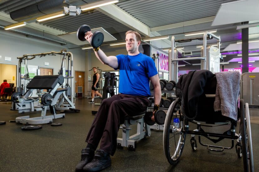 This image shows a male, out of his wheelchair and sat on a gym bench, completing a weighted arm exercise